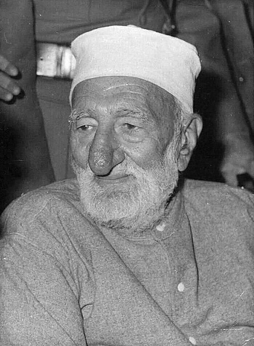 Close portrait of Bacha Khan in old age wearing a white cap, looking slightly downward, with figures standing behind him.