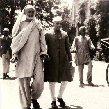 Black and white photograph: Khan Abdul Ghaffar Khan, tall and wrapped in a shawl, walking along a road beside Jawaharlal Nehru, with other figures and a horse-drawn carriage behind them.
