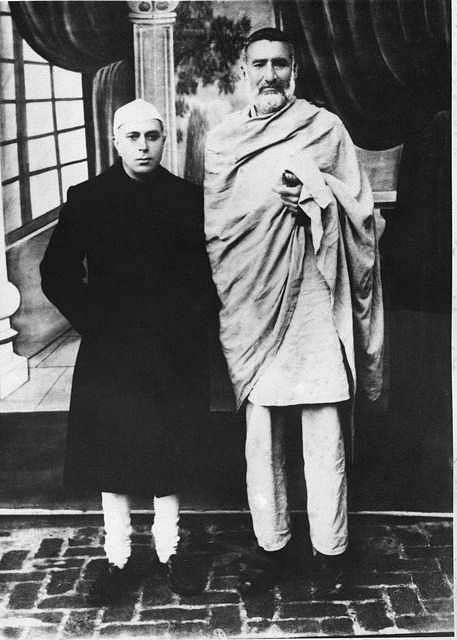Formal black and white photograph: a young Jawaharlal Nehru in a dark coat and white cap stands beside Khan Abdul Ghaffar Khan, who is much taller, wrapped in a pale shawl, on a brick-paved floor before a draped interior.