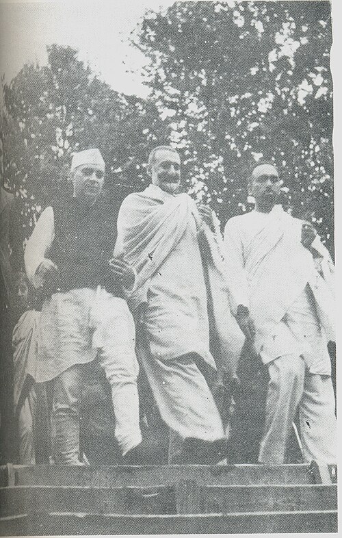 Black and white photograph of three men standing on a raised platform outdoors among trees, dressed in white, one wearing a Gandhi cap.