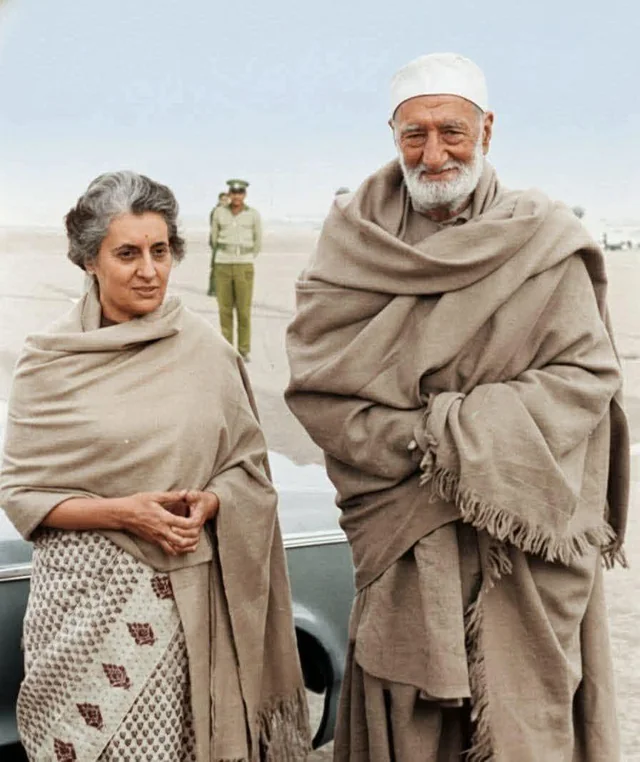 Colour photograph on an airfield: Bacha Khan in old age, tall, wrapped in a heavy fringed shawl over pale clothes and a white cap, standing beside Indira Gandhi in a shawl and printed sari.